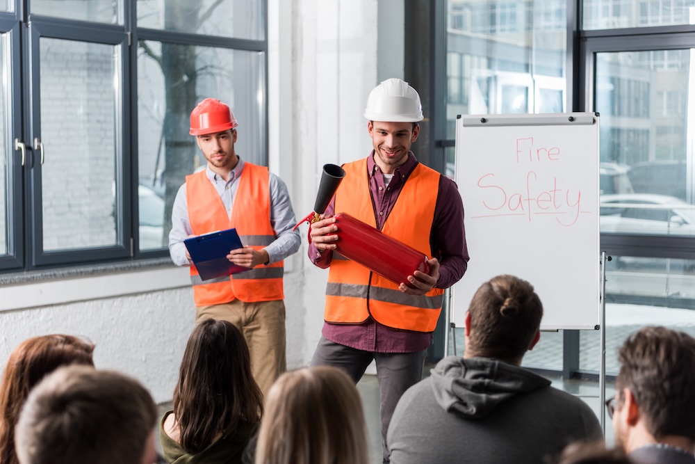 selective focus of handsome firemen in helmets standing near white board with fire safety lettering