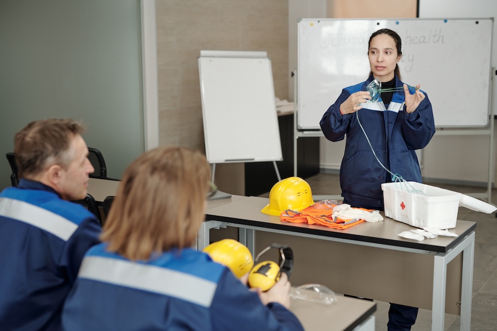 Young female worker demonstrating usage of respirator while standing by desk in front of colleagues